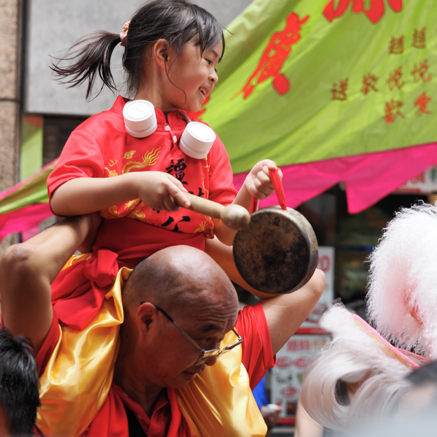 Tam Kung Birthday Parade in Shau Kei Wan – a little girl piggybacks while playing the gong. hongkong hk hkig shaukeiwan tamkung parade liondance gong